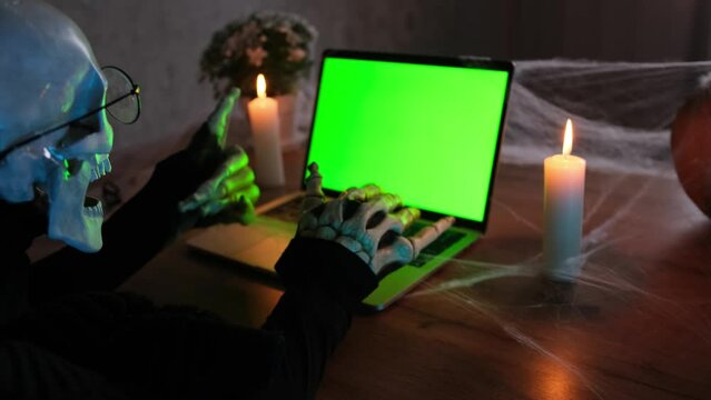 Side view man in costume death skeleton at laptop with chromakey and type scary posts and scary stories on keyboard. Celebrating holiday Halloween. Horror-loving friends to scariest Halloween party.
