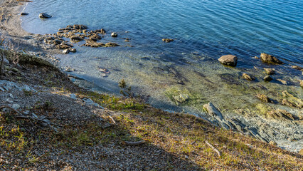 The stones are scattered in the coastal area of the lake. The bottom is visible through the clear blue water. Pebbles and broken branches on the hillside. Argentina. Tierra del Fuego. Ensenada Bay.