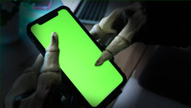 Hands in skeleton costume holding for smartphone with green screen and scrolls through social networks. Creepy Halloween party.  Blood curdling background. 