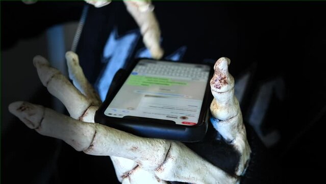 Close up, creepy bone hands in death costume uses smartphone and writes in messenger on dark background. Online predators. Creepy Halloween carnival.