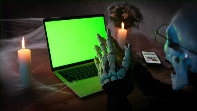 Death skeleton rubbing bone hands at laptop with chromakey and type spooky posts and scary stories, close up, side view. Happy Halloween creepy night party invitation.