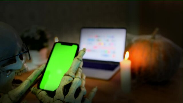 Close up, hands of skeleton holding for smartphone with chromakey scrolls through social networks and looks at photos, dark vignette. Dangers of communicating on social networks with strangers.