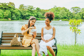Cute asian woman carries fruit basket and takes her healthy pregnant african american female friend picnic beautiful garden shady atmosphere sitting wooden bench lovingly giving gift of baby socks.