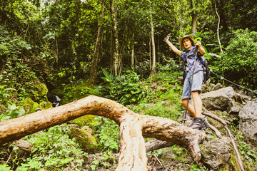 Naklejka na ściany i meble Cheerful young man enjoys hiking through the tropical forest Thailand with mountains waterfalls and lush forests riveting standing on a log looking at the beauty nature and the fresh air.