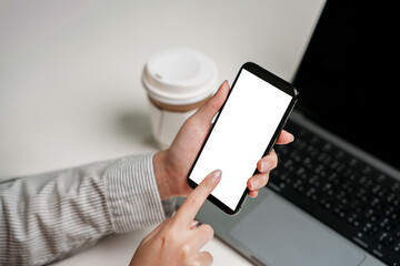 Mock up hand holding phone with finger pointing and touching on blank white screen showing online application interface social media advertising manager serving internet laptop and coffee on table