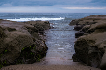 Fototapeta na wymiar 2023-08-21 ROCKY SHORELINE IN LA JOLLA WITH A WAVE ENTERING THE OPENING BETWEEN THE ROCKS WITH A NICE CLOUDY SKY