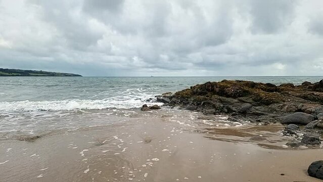 slow motion calming ocean waves washing across Anglesey island splashing over rocks on golden sandy beach, overcast morning