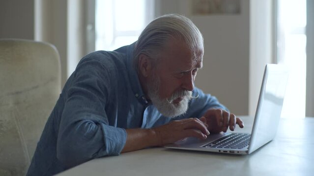 Side view of senior adult male sitting with laptop and looking at screen with concerned facial expression. Focused mature aged man looking at computer screen and squinting. Shooting in slow motion.