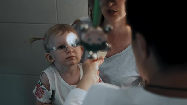 Pediatric ophthalmologist optometrist looks at eyes, checks vision of female kid. Two years old little girl child with Eye disease in glasses sits on mother's lap, doctor's appointment. Strabismus