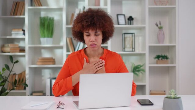 Frightened young female employee having breathing problem during panic attack at workplace. Stressed african american woman holding her chest while sitting at desk with portable laptop