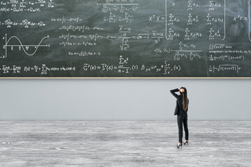 Thoughtful young european businesswoman standing in modern classroom interior with formulas on...