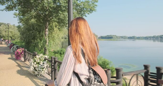 Long-haired readhead woman rides an electric scooter along the river embankment. Green trees, water, flowers