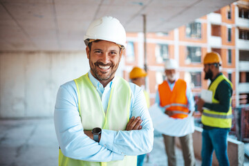 Portrait of satisfied construction site manager wearing safety vest and white helmet at...