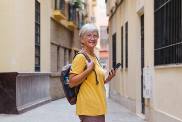 Happy senior traveler woman in yellow visiting old city of Seville in Spain, using mobile phone looking map directions