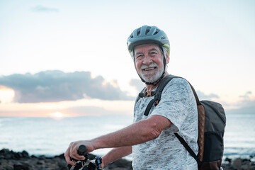 Portrait of happy active senior cyclist man at sea at sunset light with bicycle looking at camera - elderly man with helmet enjoying healthy lifestyle and freedom