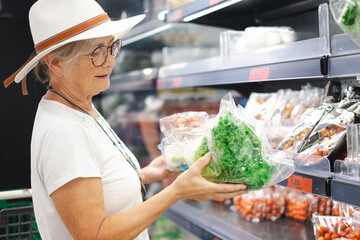 Smiling attractive senior customer woman with hat selecting vegetables at supermarket. The elderly pensioner pays attention to the increase in prices