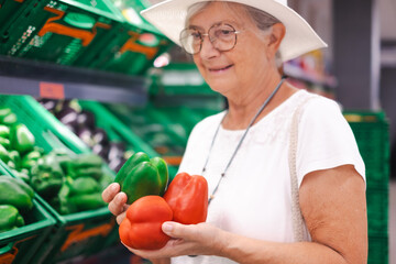 Smiling attractive senior customer woman with hat selecting vegetables at supermarket holding peppers. The elderly pensioner pays attention to the increase in prices