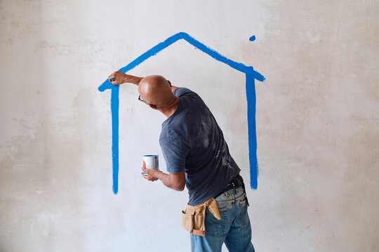Bald construction worker drawing house on wall with paintbrush at site