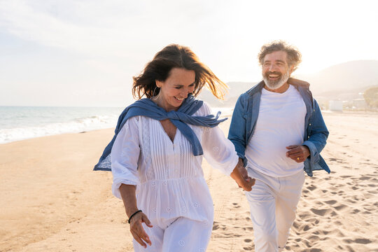 Cheerful senior woman holding hands with man and running at beach