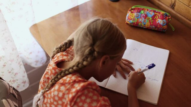 Warm scene of Little girl in orange dress making homework, top view of schoolgirl with two braids sit at table and write maths
