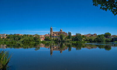 Fototapeta na wymiar Panoramic view of Salamanca Cathedral from the other side of the Tormes River with its reflection in the calm river water and all the green vegetation, clear blue sky and clean river water.