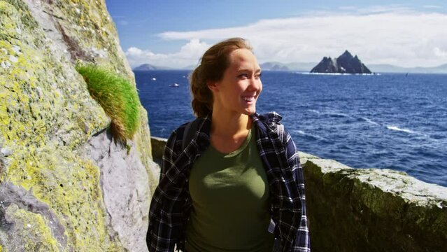 Smiling happy young woman hiking up path next to ocean with island in distance. Young explorer. 