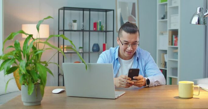 Smiling young adult asian guy student worker using mobile apps sit at home office table, happy mixed race male user enjoying browsing online social media study online on modern tech gadget