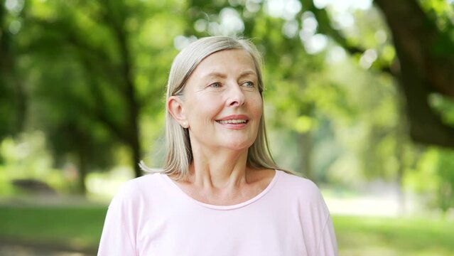 Portrait of senior active gray-haired woman standing in city park looking away. Sporty retired positive female inhales fresh air smiles. Mature old fitness happy runner enjoys being outdoors in nature