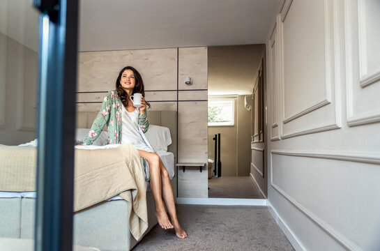 Smiling woman sitting on bed in bedroom while drinking morning coffee at home.