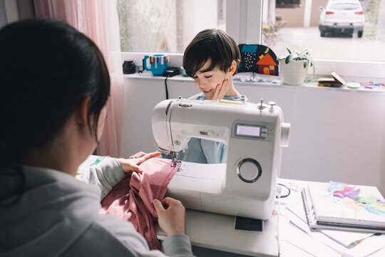 Son looking at mother sewing clothes at home