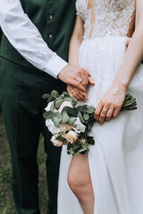 Obraz na płótnie Canvas The groom in a green suit and the bride in a white lace dress hold hands with gold rings on their fingers and a bouquet of flowers, roses at the ceremony. Wedding photography, close-up portrait.