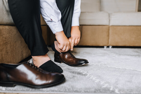 A man, the groom puts leather stylish shiny brown shoes on his feet in socks in the morning, sitting on the sofa. Photography, businessman portrait, business.