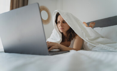 Young sad woman lying on the bed in the morning and looking at the laptop screen hiding under the blanket