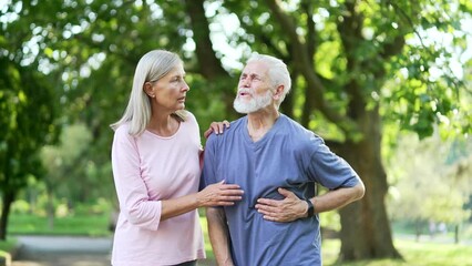 Sick senior gray haired man having a heart attack during training in an urban city park. Mature old wife supports her husband. The male holds his hand to his chest. He needs urgent medical attention