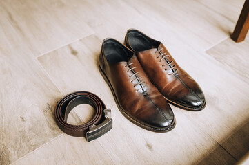 Men's brown leather shoes and a black belt lie on the floor. Close-up photo, top view.