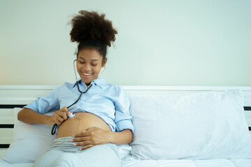 Happy cheerful American - African black ethnicity pregnant woman sitting on the bed in bedroom and listening a baby voice inside her belly on stethoscope.