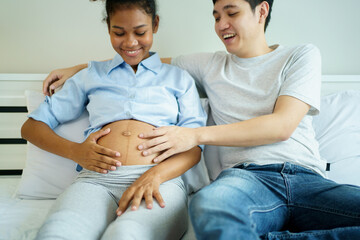 American - African black ethnicity woman and her Asian husband relaxing together on the bed in bedroom and a man tenderly touching on his wife belly. Husband taking care of his pregnant wife.