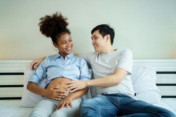 American - African black ethnicity woman and her Asian husband relaxing together on the bed in bedroom and a man tenderly touching on his wife belly. Husband taking care of his pregnant wife.