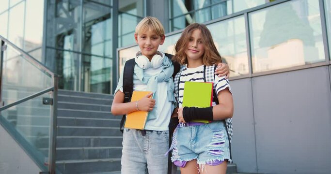 Portrait of cheerful smiling two caucasian elementary school children standing posing on the stairs near school in outdoor looking at camera happy after school reopen. Back to school concept