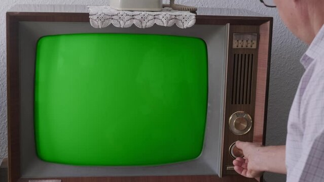 elderly man turns on and watches old retro analog TV with blank green screen for designer, entertainment, leisure essential of life older individuals, Technology 1960-1970, mockup, template for video
