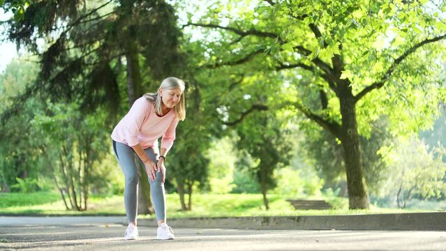 Senior gray haired woman suffers from painful severe knee joint ache during a run in city park. Mature old female massaging stretching trauma injury while jogging outdoors. Leg muscle cramp calf sport