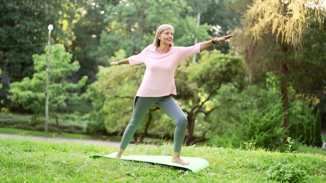 Senior active gray haired woman does gymnastics while standing on a mat in an urban city park. Mature old fitness female doing yoga outside. Elderly sportswoman performs muscle stretching exercises