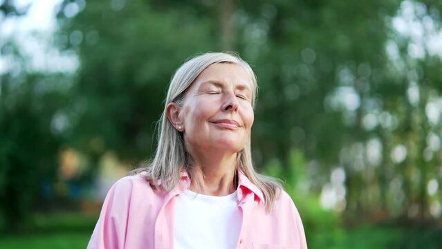 Portrait of senior adult gray haired woman relaxing with closed eyes standing in urban city park. Retired female breathing deeply smiling looking at camera. Mature old pensioner enjoys being in nature