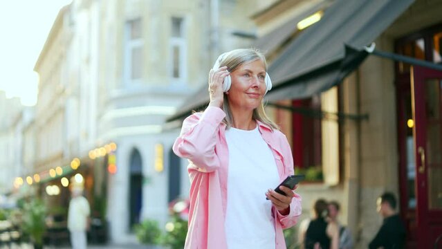 Senior gray haired woman walking with headphones mobile phone enjoying listening to music outdoors on city street. Happy mature old female in a shirt includes playlist and relaxing at leisure outside