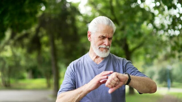 Close up. Senior active gray haired bearded runner looking smart watch standing in city park. Mature old male using fitness tracker and checking result. Elderly sport man on training workout in nature