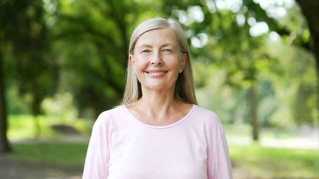Portrait of senior active gray-haired woman standing in city park. Sporty retired positive female looking at camera and smiling. Mature old fitness happy runner posing in nature. Headshot of pensioner
