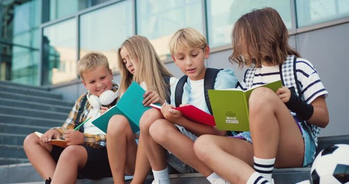 Happy caucasian school friends talking and doing homework, having fun during break sitting on stairs at school building background, outdoor. Back to school concept