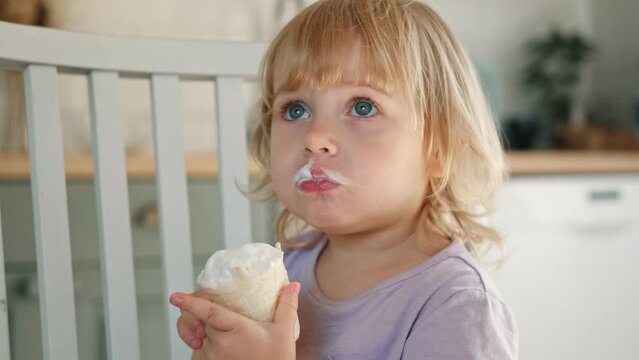 Baby girl enjoying ice cream. Pretty little toddler eating an ice-cream indoors, at home. Dining room background. Small child eats plombir and cream messy on her mouth. Cute kid with tasty sweet food.