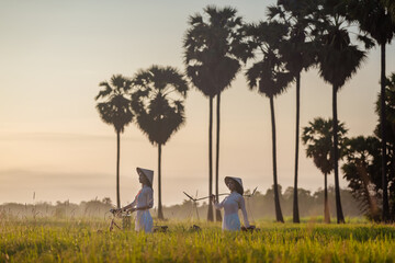 Beautiful Vietnamese woman wearing traditional dress with bicycle and Carrying things walk pass rice field on sunrise time ,lifestyle of farmer in Vietnam with sugar palm tree background.