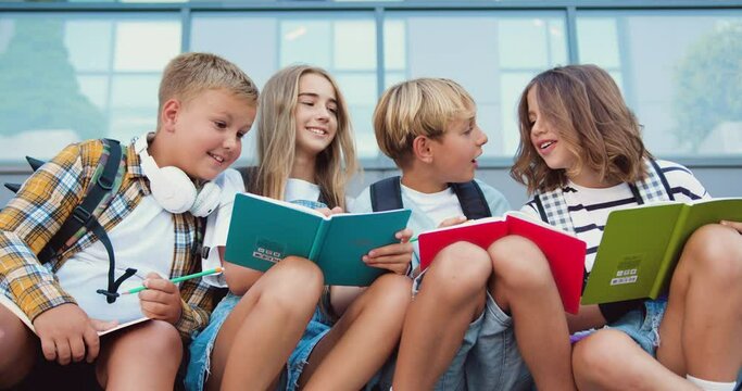 Close up of four caucasian school friends talking and doing homework, having fun during break sitting on stairs at school building background, outdoor. Back to school concept